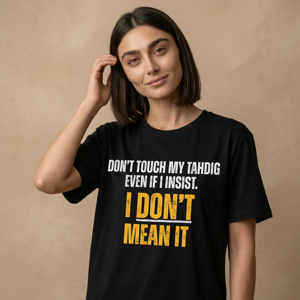 A woman with dark shoulder-length hair wearing a black graphic t-shirt with the text 'DON'T TOUCH MY TAHDIG EVEN IF I INSIST. I DON'T MEAN IT' in white and yellow, posed against a textured beige background.