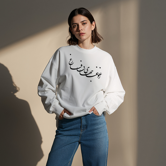 A person wearing a white sweatshirt with a Persian calligraphy text and blue jeans standing against a plain wall.